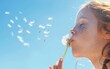 Young girl blowing a dandelion under a clear blue sky.