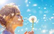 Young girl blowing dandelion seeds into a clear blue sky.