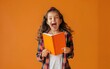 Young girl in plaid shirt, excitedly holding an orange notebook, orange background.