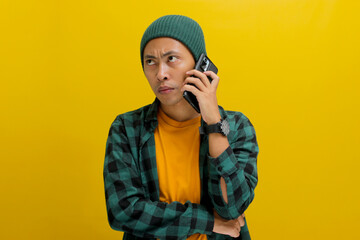 Wall Mural - Annoyed Asian man, dressed in beanie hat and casual clothes, engages in a phone conversation, looking away at an empty copy space with confused and displeased expression, isolated on yellow background
