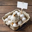 A baby chick stands in a dozen eggs in a cardboard egg carton with a blank sign attached to a stick behind it