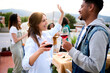 © CarlosBarquero - Excited multiracial young couple celebrating rooftop party drinking red wine. Friends dancing at sunset date weekend gathering. Cheerful gen z people enjoying romantic leisure outdoors.