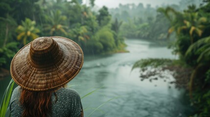 A woman sporting a traditional bamboo hat gazes out at the river and lush rainforest from her vantage point capturing the essence of the rainy season on a tropical island This picturesque s