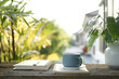 Blue coffee cup on wooden tray and notebook and Philodendron plant on wooden table under sunlight