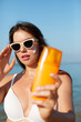 Beautiful young woman with sun cream on face. Girl holding sunscreen bottle on the beach and applying moisturizing lotion on skin.Skin care. Sun protection. Suntan