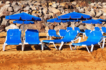 Wall Mural - Sunbeds and umbrellas on the sandy beach with rocks. Blue sun loungers