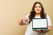 Cute Latin plus-size woman in a studio pointing at a blank laptop screen