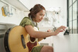 Young Asian woman enjoying a guitar session while following a song on her smartphone in a cozy kitchen