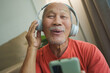 Joyful elderly Asian man enjoying music with new headphones, holding a smartphone at home