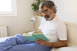 Mature man with glasses relaxing in bed, reading a book wearing pyjamas.
