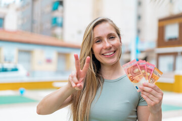 Wall Mural - Young blonde woman taking a lot of money at outdoors smiling and showing victory sign