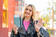Young pretty blonde woman holding a passport at outdoors saluting with hand with happy expression