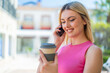 Young pretty blonde woman at outdoors using mobile phone and holding a coffee with happy expression