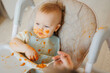 Happy messy baby boy eating pasta with tomato enjoys food sits In high chair, light background