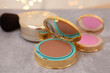 Face bronzer and other cosmetic products on grey textured table against blurred lights, closeup