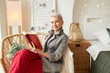 Indoor portrait of elegant stylish elderly business lady in denim jacket and red pants having rest sitting in big cozy wicker chair with book in hands, enjoying leisure time in her country house