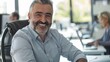 © iuricazac - Smiling man with gray beard and hair wearing blue shirt sitting in office chair looking at camera with blurred background of office environment.