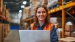 © iuricazac - Smiling woman in orange vest working on laptop in warehouse.