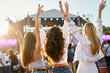 © artiemedvedev - Back view of three young women at beach music festival, arms raised, dancing, enjoying summer sunset party vibe. Friends celebrate, fun outdoor event, casual fashion, stage lights in background.