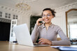 © muse studio - A cheerful young woman engages in a phone conversation while working on a laptop in an elegant, traditional office setting.