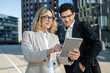 © muse studio - Two business professionals, a woman and a man, review content on a tablet together, smiling, outdoors in an urban setting.