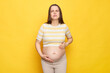 Sick Caucasian pregnant woman in casual clothing posing isolated over yellow background feeling discomfort and pin in her belly grimacing touching her tummy