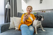 Elderly Caucasian woman sitting with her pet French bulldog while sitting on the sofa at home and using smartphone