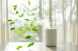 A modern white air purifier on a table, emitting steam with green leaves swirling around, against a large window with a sunny backdrop. Shallow depth of field