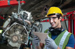 A young male mechanic wearing a hard hat and safety glasses smiles while using a tablet in an automotive repair shop.