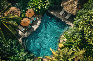 An aerial view of the pool and loungers at a luxury hotel in Bali, surrounded by lush greenery. The oval shaped pool has two sunshades over it, surrounded by palm trees and other tropical plants.