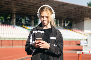 Teenager girl uses a smartphone and listens to music on headphones at the stadium, outdoor workout concept.