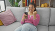 © Krakenimages.com - A joyful hispanic woman celebrates a victory while looking at her smartphone, surrounded by cozy home decor.