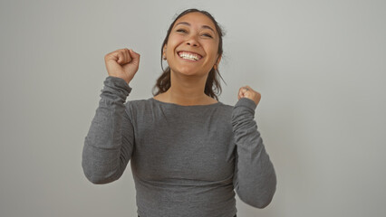 Poster - Joyful young hispanic woman celebrating with clenched fists against a plain white wall in a casual gray top.