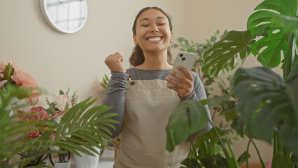 Sticker - A joyful woman celebrates with a smartphone in a lush flower shop surrounded by foliage and blooms.