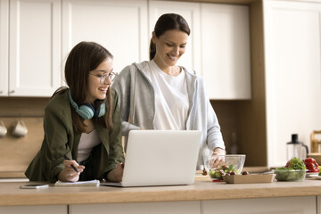 Happy smart teen school girl and positive young mom studying online and cooking salad for dinner at kitchen table, using laptop, chopping vegetables, talking, laughing, having fun