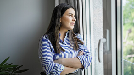 Poster - Young beautiful hispanic woman standing smiling looking through the window at home