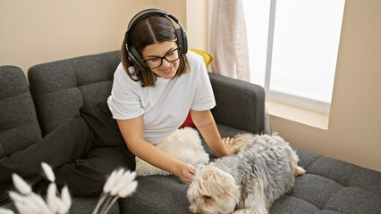 Canvas Print - A smiling hispanic woman in headphones pets a dog on a couch in a cozy living room setting.