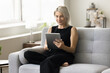 © fizkes - Positive blonde mature woman using online application on tablet computer at home, relaxing on couch, smiling, reading book on Internet, enjoying wireless Internet technology on digital device
