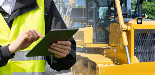 Poster - Worker in a helmet with a digital tablet on the background of construction machine..