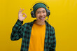 Smiling Asian man in a beanie and casual clothes gives an okay sign while listening to music or a podcast on his headphones. Isolated on a yellow background.
