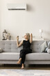 © fizkes - Happy relaxed middle aged woman starting air conditioner from comfortable sofa, resting on couch at home, using conditioning cooling ventilation split system with equipment on wall