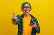 Smart young Asian man, sporting eyeglasses and dressed in a beanie hat and casual shirt, gives a thumbs up gesture while holding a phone, expressing approval, agreement, and offering a positive review