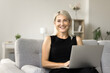 © fizkes - Happy beautiful blonde mature freelance business woman sitting on couch with laptop, looking at camera with toothy smile, laughing, posing for home portrait, enjoying online communication