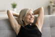 Happy dreamy pretty mature woman resting on comfortable sofa, relaxing at home, breathing fresh cool air, looking away with toothy smile, thinking, dreaming on calm lazy retirement