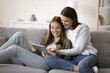 Happy mother and teenager kid girl using online application for shopping together, resting on sofa, holding digital tablet computer, smiling, laughing, enjoying domestic Internet communication