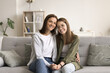 Happy pretty teenage kid girl and beautiful mom sitting together on sofa, looking at camera, smiling. Mother holding daughters hand, hugging teen child with care, love. Home family portrait