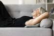 Cheerful calm mature woman resting on sofa with happy face, smiling, laughing, thinking, dreaming on good future plans, breathing cool fresh air, looking away, relaxing at home