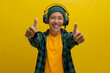 Asian man in a beanie and casual clothes smiles in approval while listening to music on his headphones. He gives a thumbs up gesture, seemingly enjoying the music. Isolated on a yellow background.