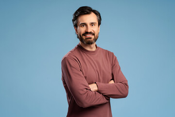 Wall Mural - Positive bearded young man with arms crossed, looking at camera posing in studio, isolated on blue