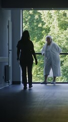 Wall Mural - Clinic corridor: Doctors and professional medics walk. Nurse with papers comes to elderly female patient standing near window. Medical staff and patients in dark hospital or medical center hallway.